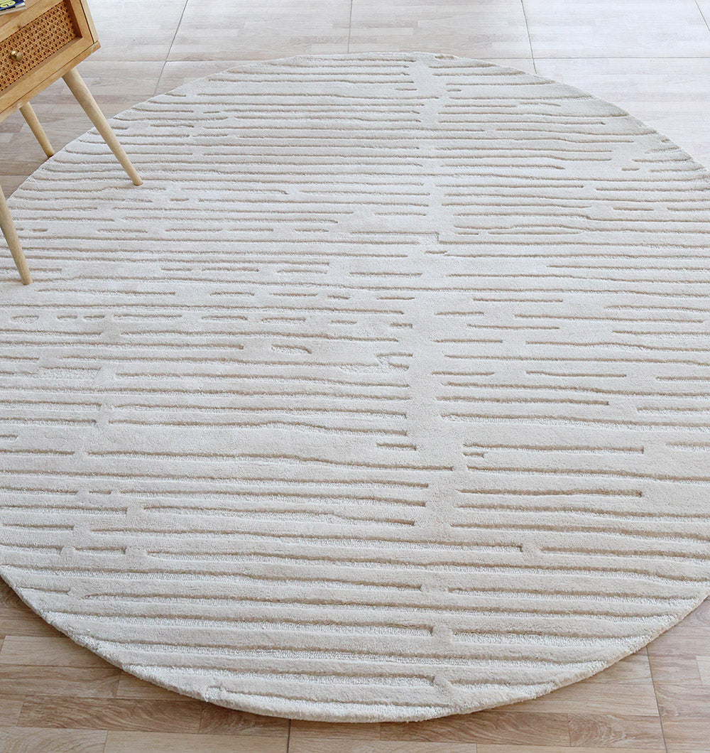 The RAMSHA HOME Home Decor Handmade Area Rug - HTV 17, a round, cream-colored rug with a subtle linear pattern, sits on a light wood floor beside a wooden chair with a woven seat.