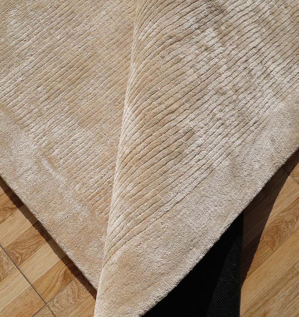 A close-up of the RAMSHA HOME Beige Color Handmade Rug - HTV 18 (200x300 cm), showing its ribbed texture and a lifted corner that reveals a black non-slip mat beneath on a wooden floor.