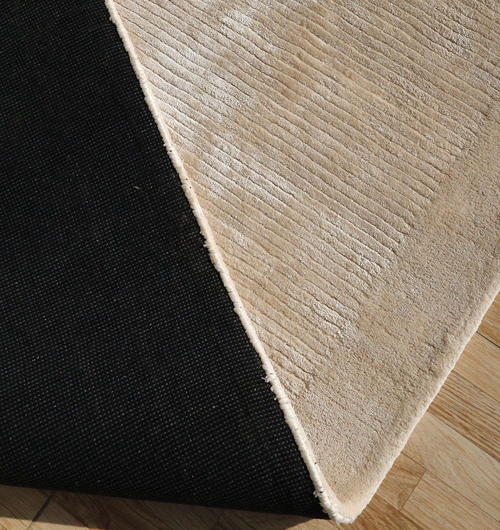 The RAMSHA HOME Beige Color Handmade Rug - HTV 18 (200x300 cm) features a textured surface and is shown partially flipped to reveal its black non-slip backing, displayed on a light wood floor.