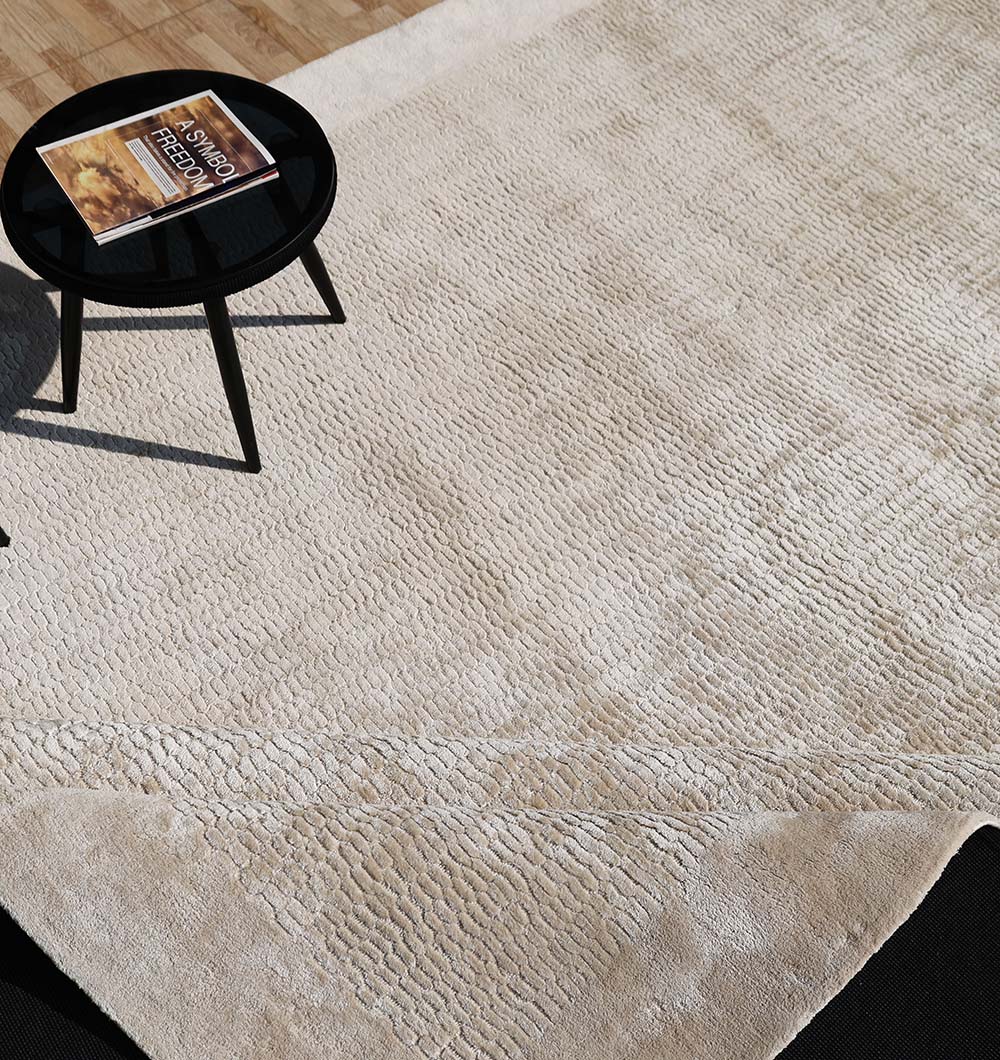 A RAMSHA HOME Beige Color Handmade Area Rug - HTV 20 (300x400 cm) covers the wooden floor. On the rug, a small round black table holds two stacked books, with sunlight casting gentle shadows across the scene.