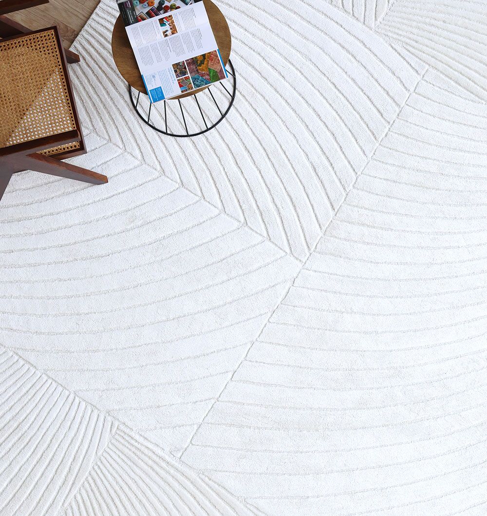 A modern living space featuring the RAMSHA HOME Viscose & Cotton Handmade Area Rug - HTV 22 (300X400 cm) with geometric lines, a wooden chair with a woven seat, and a round side table holding an open magazine and a blue item.
