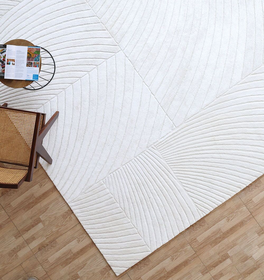 A RAMSHA HOME Viscose & Cotton Handmade Area Rug - HTV 22 (300x400 cm) with geometric curves covers part of a wood floor, topped with a round side table holding a magazine and a wooden chair with a woven seat.