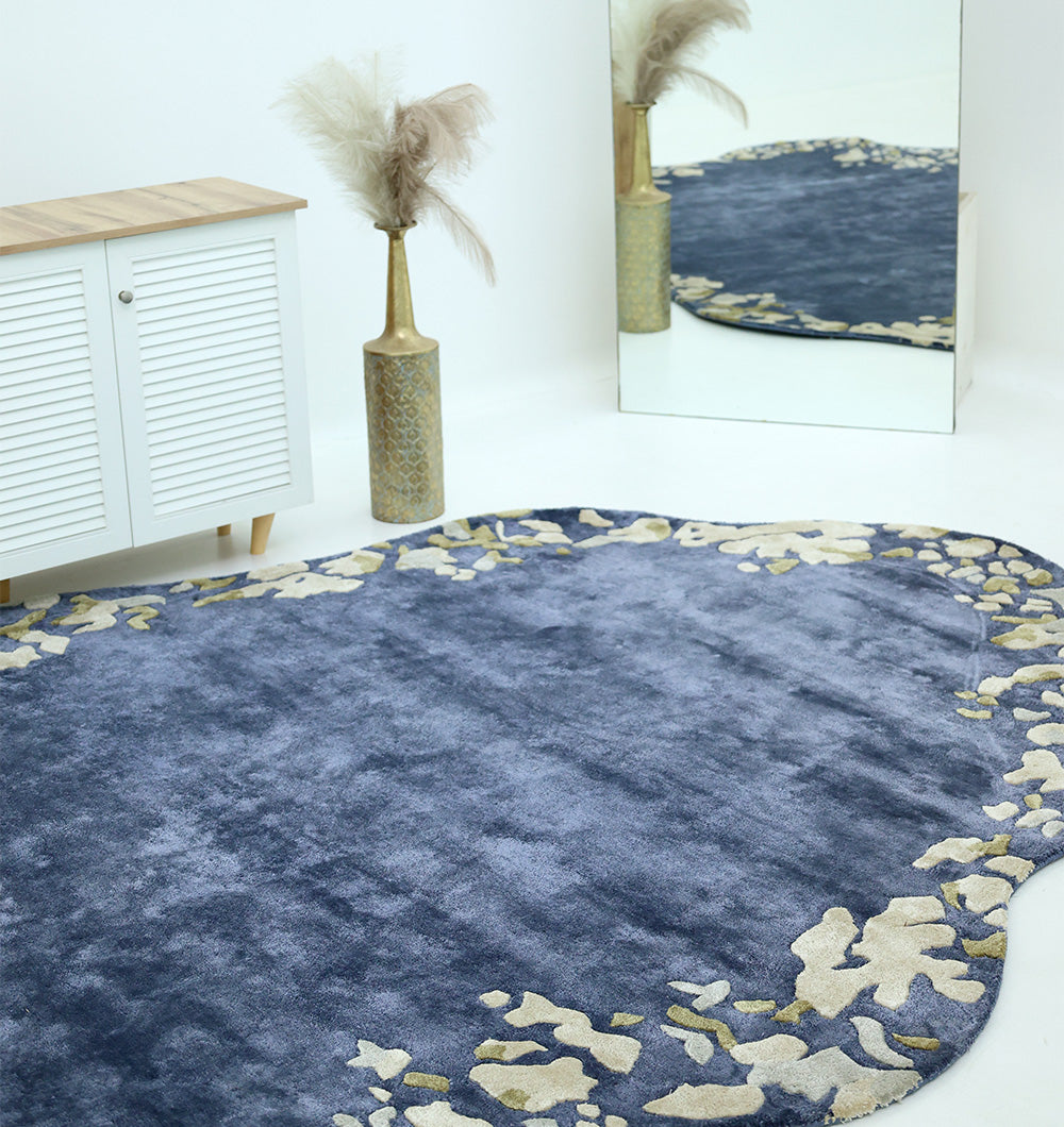 The RAMSHA HOME Navy Blue Viscose Shaped Handmade Carpet 200x300 (HTV-23), featuring abstract cream edge patterns, is placed on a white floor next to a wooden cabinet, pampas grass vase, and a large mirror.