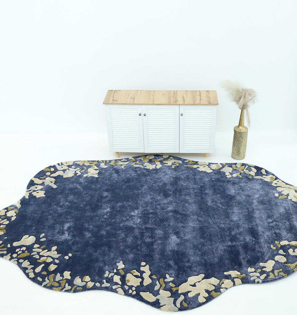 The RAMSHA HOME Navy Blue Viscose Shaped Handmade Carpet 200x300 (HTV-23), featuring beige abstract patterns, lies on a white floor in front of a small white cabinet with a wooden top and a tall vase with pampas grass beside it.
