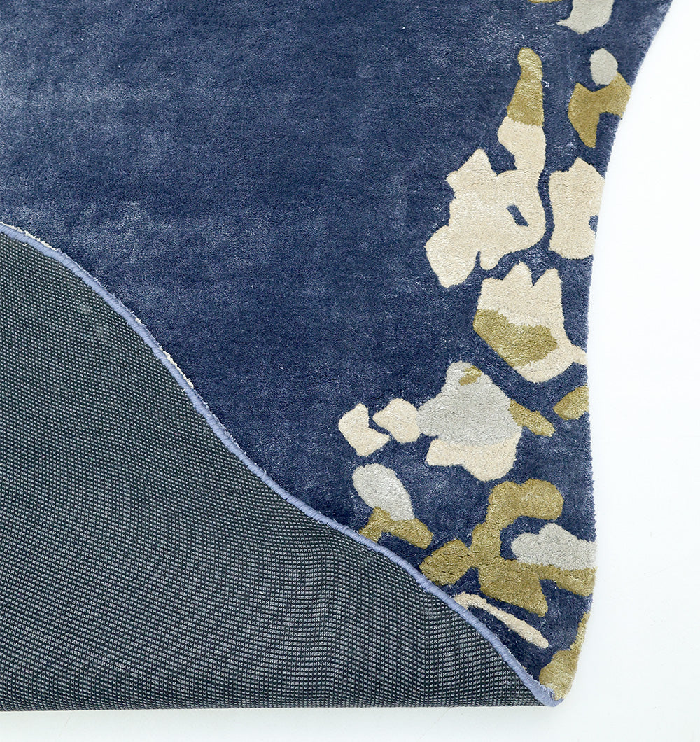 A close-up shows the corner of the RAMSHA HOME Navy Blue Viscose Shaped Handmade Carpet 200x300 - HTV-23, featuring abstract beige and olive green designs, partially flipped to display its gray non-slip backing.
