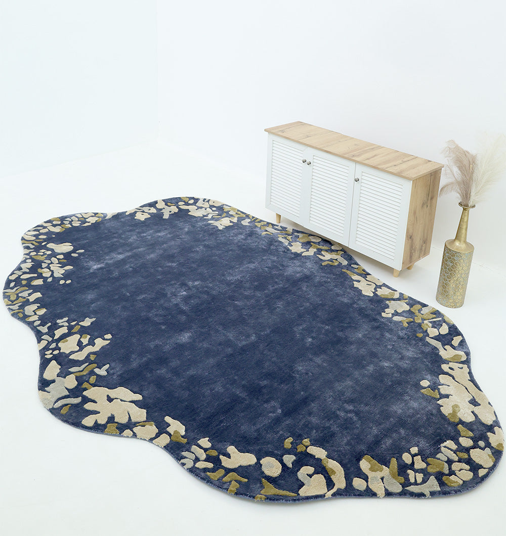The RAMSHA HOME Navy Blue Viscose Shaped Handmade Carpet 200x300 HTV-23, with abstract cream and gold accents, decorates a minimalist white room featuring a wooden cabinet and a vase of pampas grass.