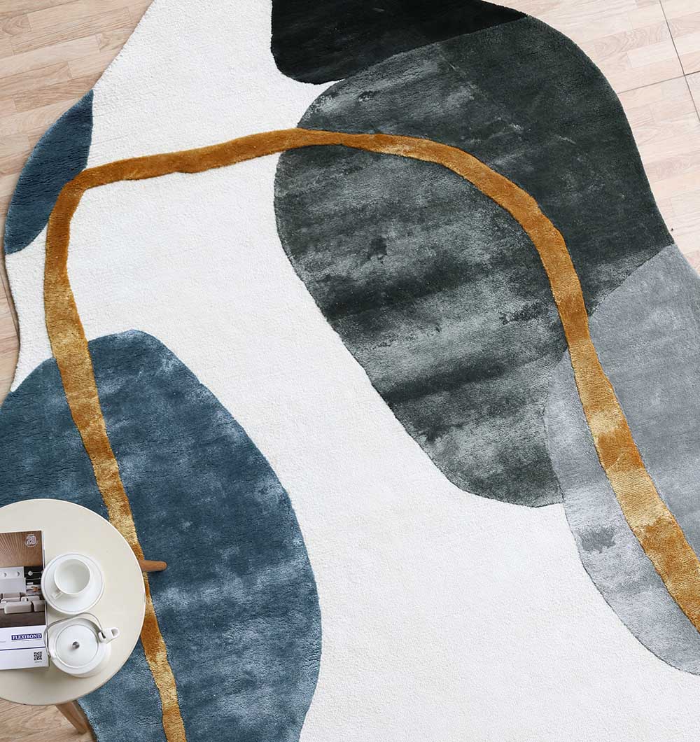 The RAMSHA HOME Handmade Modern Shaped Rug For Home Decor HTV 24 (200 x 300 cm) features an off-white base with bold blue, dark green shapes, and a curving mustard yellow line. Styled as an irregular area rug under a round table with tea and magazines.