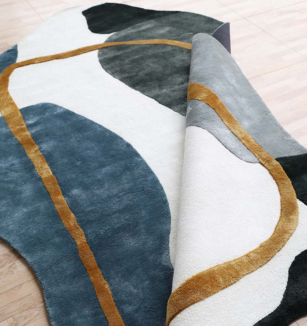 The Handmade Modern Shaped Rug For Home Decor HTV 24 by RAMSHA HOME (200 x 300 cm) features abstract blue, white, gray, and dark green shapes with curved mustard yellow lines; one corner is folded to show the same tencel pattern below.