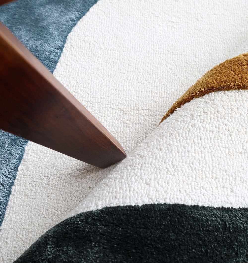 Close-up of the RAMSHA HOME Handmade Modern Shaped Rug For Home Decor HTV 24 (200 x 300 cm), featuring white, blue, green, and mustard sections. A wooden furniture leg sits on the irregular rug, with one edge slightly lifted.