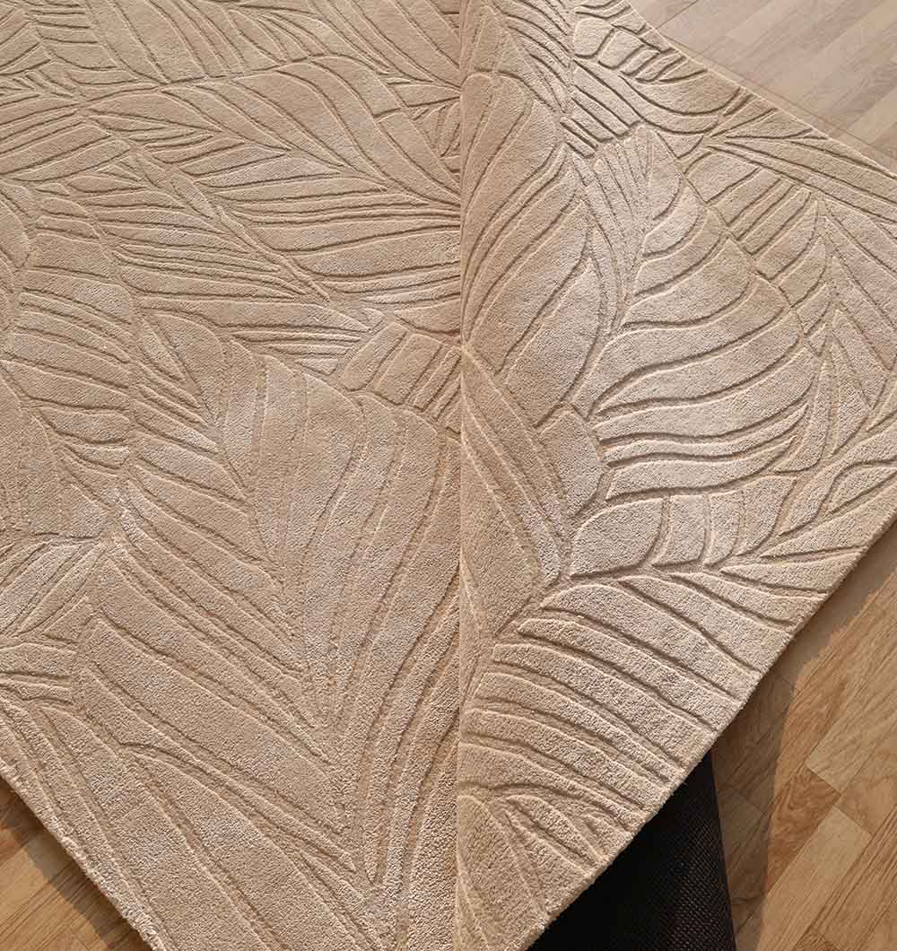 A close-up of the RAMSHA HOME Rectangle Shape Beige Color Handmade Area Rug - HTV 25 (200X300 cm), featuring a textured leaf pattern, shown partially folded to reveal its underside on a wooden floor.