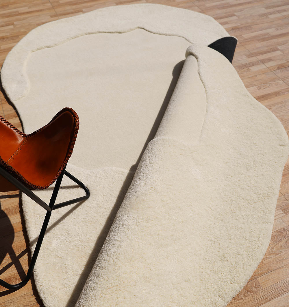 The RAMSHA HOME Handmade Shape White Color Area Rug - HTW 101 B is partially rolled back on a light wood floor, with a brown leather chair featuring a black metal frame visible in the left corner of the image.