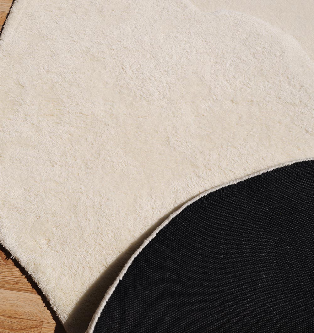 A close-up of the RAMSHA HOME Handmade Shape White Color Area Rug - HTW 101 B on a wooden floor, showing one corner flipped to display its black non-slip backing and unique textured surface.