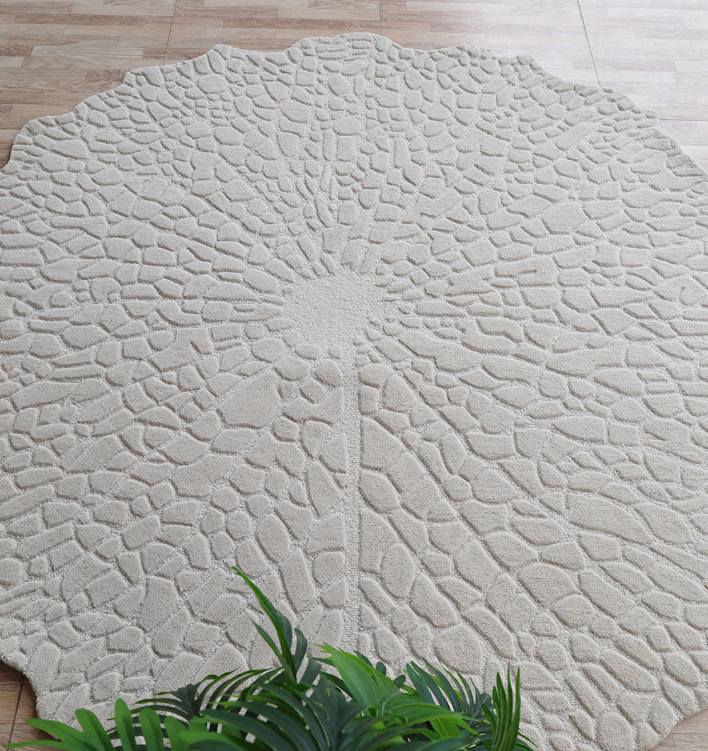 The Handmade Beige Color Area Rug HTW 103 by RAMSHA HOME, featuring a dandelion-inspired pattern and cream textured wool, lies on a wooden floor with green plant leaves in the foreground. Its petal-like segments create a neutral statement piece.