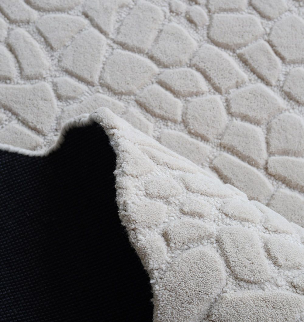 A close-up of the RAMSHA HOME Handmade Beige Color Area Rug HTW 103 shows its raised, irregular stone pattern; one corner of the textured wool rug is folded over to reveal a black fabric backing.