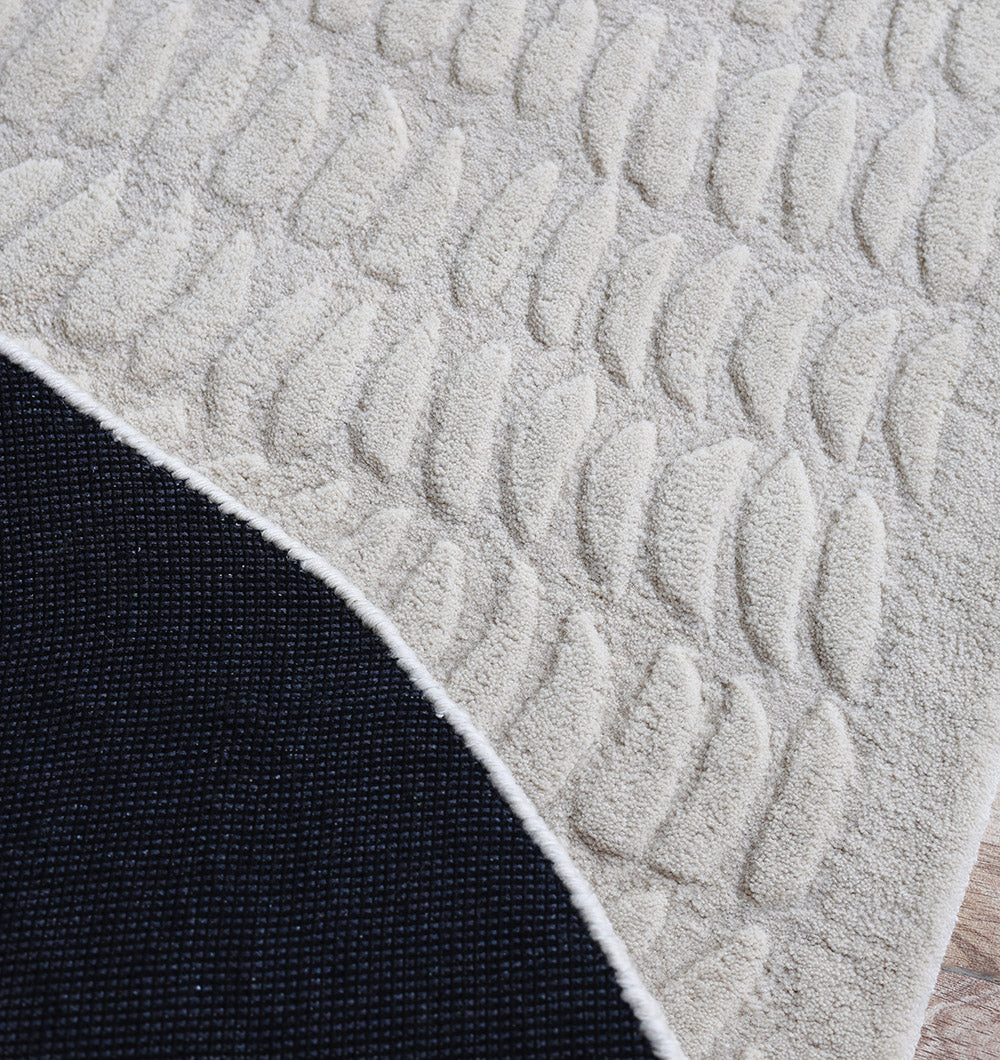 A close-up of the RAMSHA HOME Beige Color Shape Handmade Rug HTW 104 shows its modern textured leaf pattern and cream tone. The flipped corner reveals a dark non-slip backing, all set on a wooden floor.