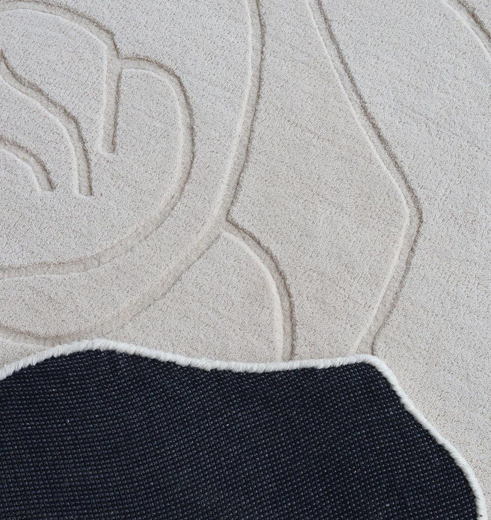 A close-up of the RAMSHA HOME Handmade Rose Shape Rug HTW 106, featuring textured cream tones with abstract raised patterns, partially covered by a dark woven fabric accented with white trim.