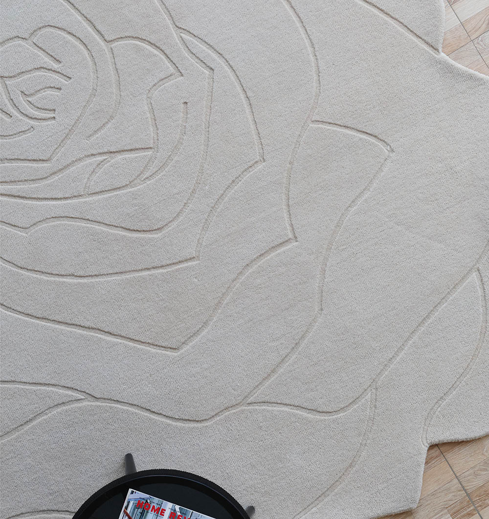 The RAMSHA HOME Handmade Rose Shape Rug HTW 106, in beige with a large embossed rose design, lies on a light wooden floor. A round black table with magazines appears partially in the bottom left corner.