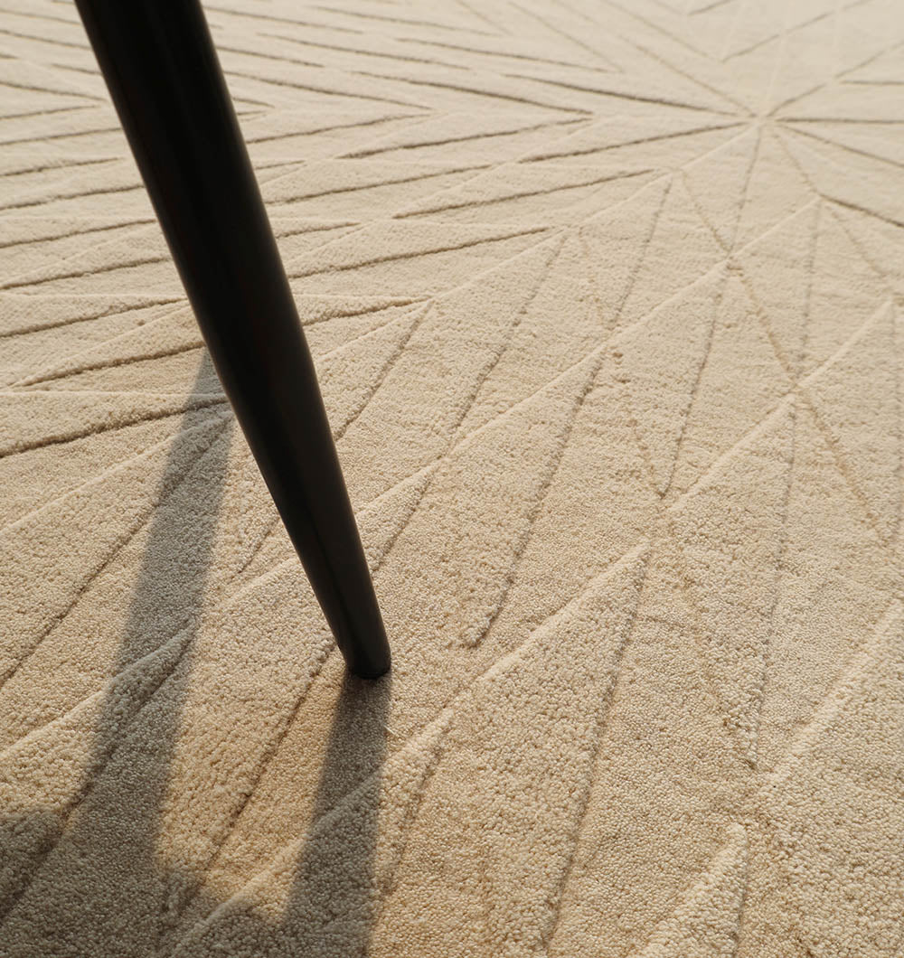 A close-up of the RAMSHA HOME Beige Handmade Round Rug HTW 107 (200cm) shows its geometric patterns, with the shadow and leg of a black table or chair visible.