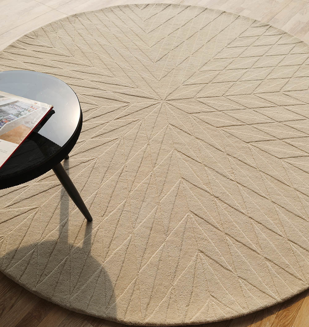 The RAMSHA HOME Beige Handmade Round Rug HTW 107 (200cm) showcases artisanal quality with its geometric starburst design, pictured on a wooden floor next to a black round side table holding an open magazine.