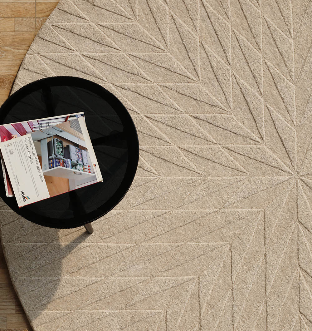 The RAMSHA HOME Beige Handmade Round Rug HTW 107 (200cm), with a geometric starburst pattern and raised lines, sits on a light wood floor beneath a round black coffee table with magazines.