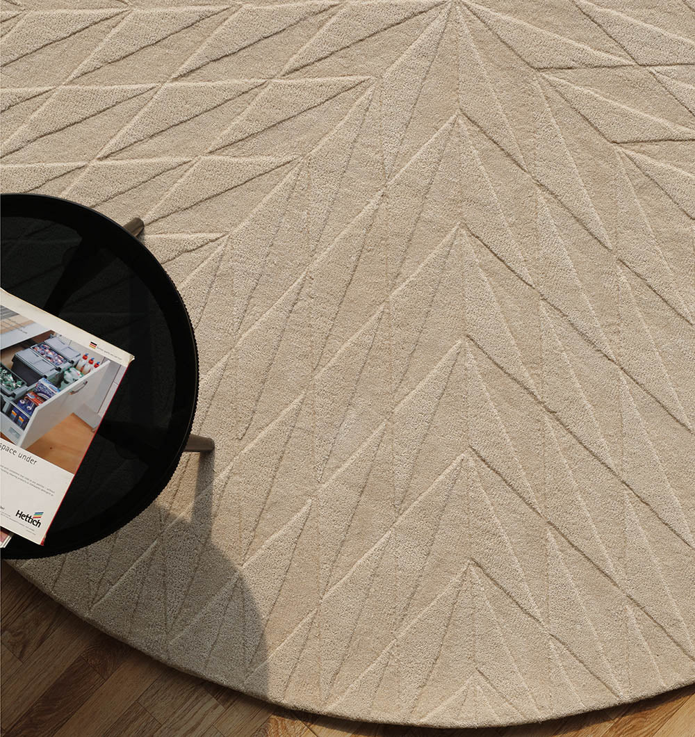 The RAMSHA HOME Beige Handmade Round Rug HTW 107 (200cm) features a geometric arrow-like texture and artisanal craftsmanship, shown on a wooden floor with a small black round table holding magazines in the corner.