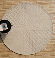 The RAMSHA HOME Beige Handmade Round Rug HTW 107 (200cm) with a geometric starburst pattern lies on a light brown wooden floor, partially under a small black side table holding magazines.
