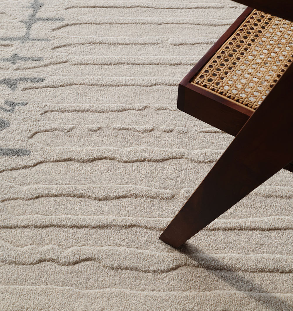 A close-up of the RAMSHA HOME Irregular Shape Handmade Rug HTW 108, featuring textured beige wool and raised patterns, beside the leg and woven cane seat of a wooden chair.