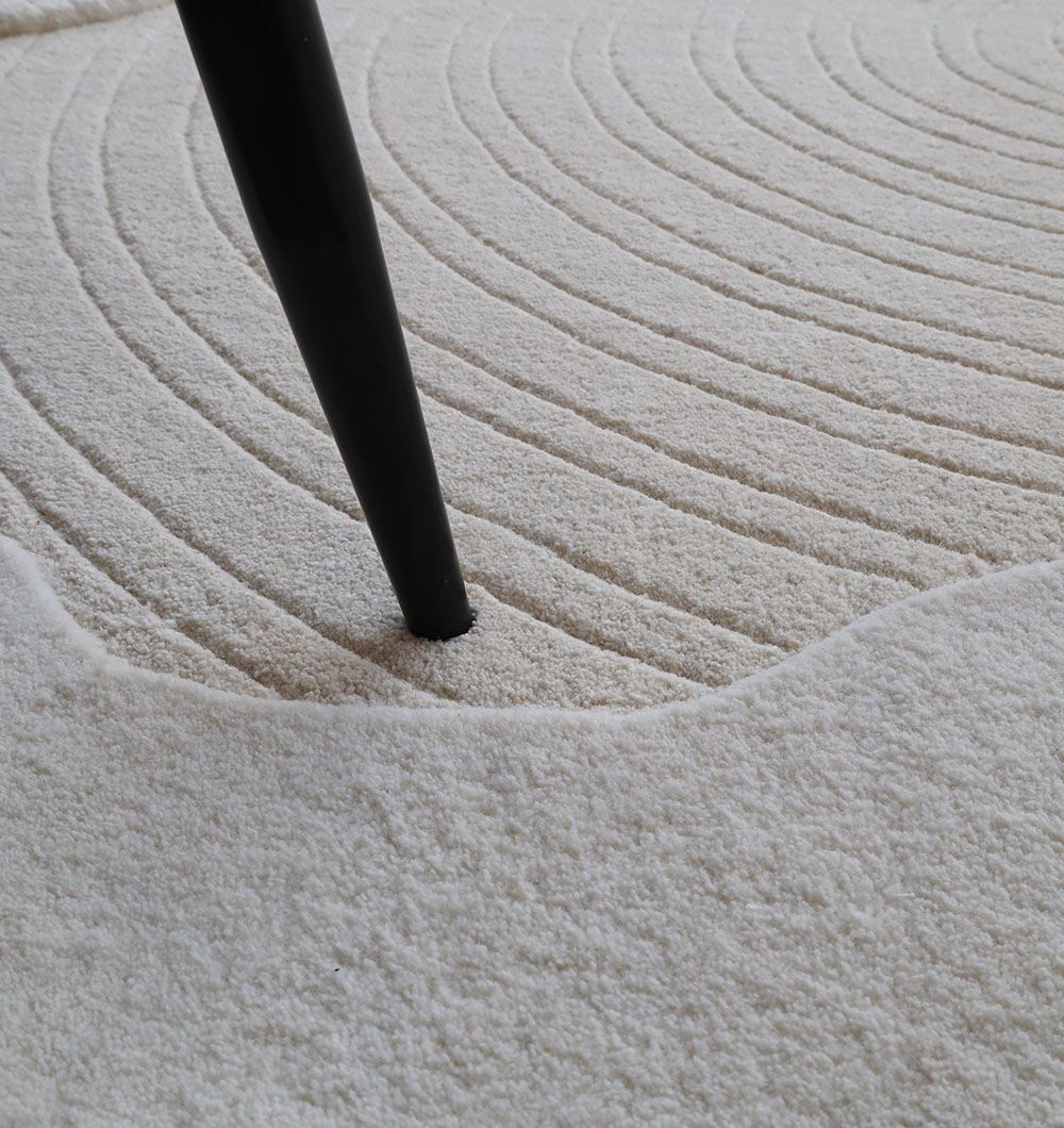 A close-up of a black table leg on the RAMSHA HOME Multi Color Handmade Area Rug - HTW 109 (183x200 Cm), featuring textured cream wool with concentric curved lines and wavy patterns for a calming, zen-inspired look.