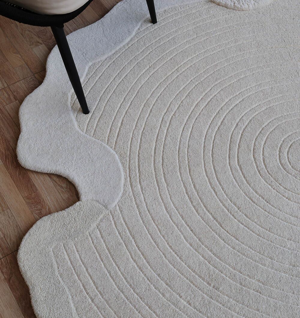 The RAMSHA HOME Multi Color Handmade Area Rug - HTW 109 (183x200 Cm), a modern hand-tufted wool rug with concentric circles and wavy edges, lies on a wooden floor, partially beneath a chair with black legs and a light seat.