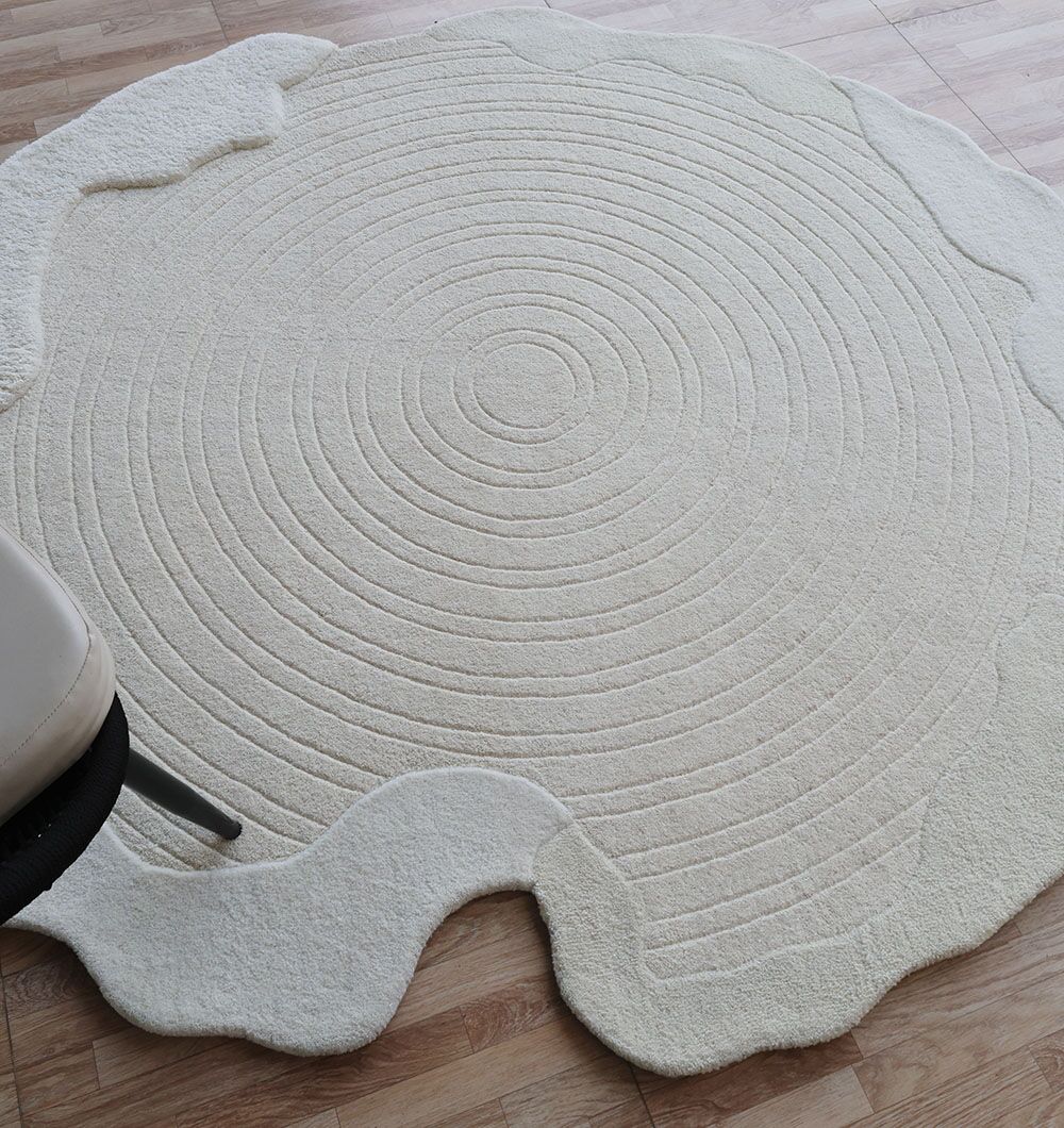 The RAMSHA HOME Multi Color Handmade Area Rug - HTW 109 (183x200 Cm) features concentric circles and wavy edges, displayed on a wooden floor with a light chair at its edge, creating a modern neutral accent.