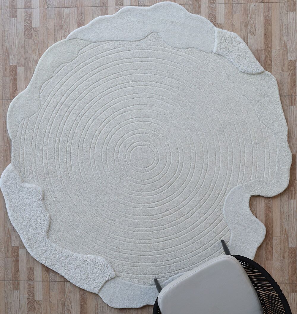 The RAMSHA HOME Multi Color Handmade Area Rug - HTW 109 (183x200 cm) features concentric ring patterns in cream, resembling tree rings, on a wood-patterned floor; a light-cushioned chair at the corner enhances its modern, inviting style.