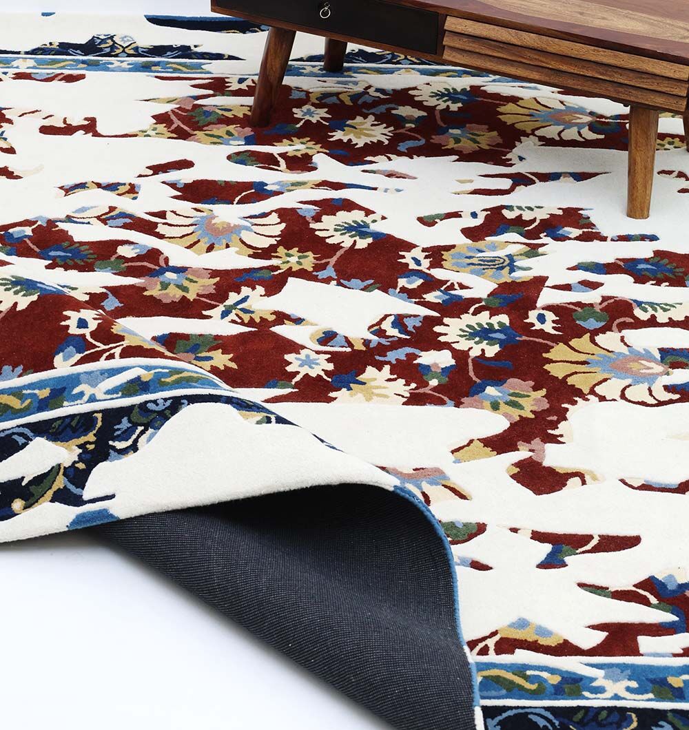 The RAMSHA HOME Multi Color Handmade Area Rug - HTW 111 (300X400 cm) with a floral design in red, blue, and gold lies partially folded near a wooden table with tapered legs on a white floor.