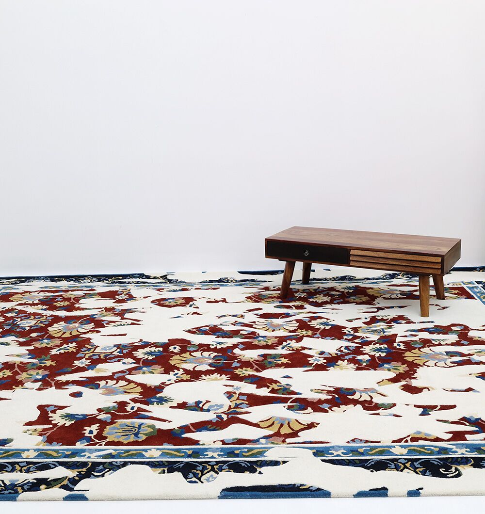 A low wooden table with a drawer sits on the RAMSHA HOME Multi Color Handmade Area Rug - HTW 111 (300X400 cm), featuring ornate red, blue, and beige patterns, set against a plain white wall and floor.