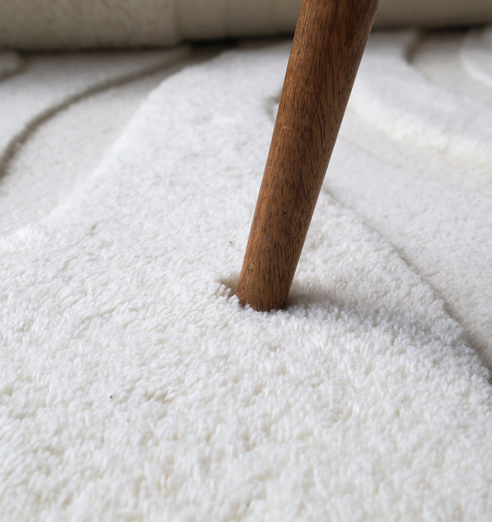 A wooden furniture leg presses into the Luxury Off White Handmade Area Rug - HTW 114 (200x300 cm) by RAMSHA HOME, showing an indentation in its plush, looped fibers and emphasizing the rug’s cozy, neutral appearance.