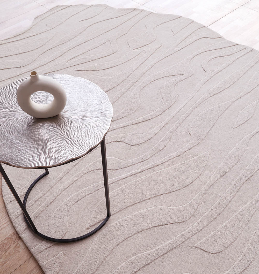 A round metallic side table with a textured surface holds a circular vase, set on the Irregular Shape Beige Color Handmade Rug HTW 116 by RAMSHA HOME, featuring abstract wavy raised patterns in a minimalist room.