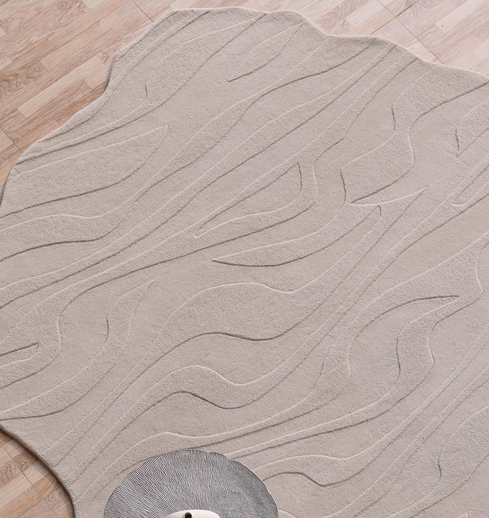 The RAMSHA HOME Irregular Shape Beige Color Handmade Rug HTW 116, featuring raised wavy patterns, lies on a light wood floor. A glimpse of a textured, circular decorative object appears in the lower left corner.