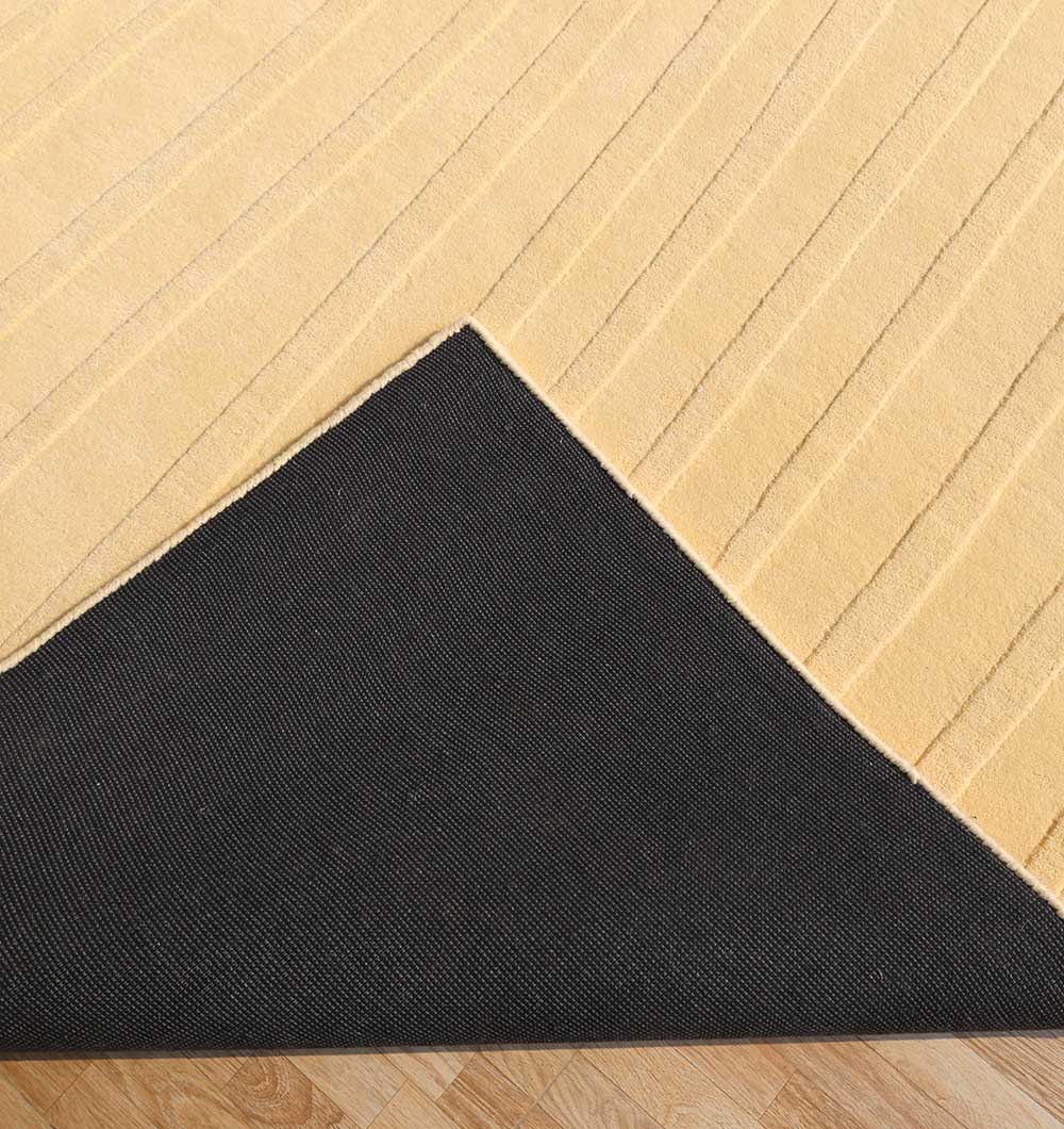 A close-up shows the RAMSHA HOME Minimal Wool Handmade Carpet 300x400 Yellow – HTW-117 in beige with subtle linear patterns, one corner flipped to reveal its black non-slip backing, placed on a light wood floor.