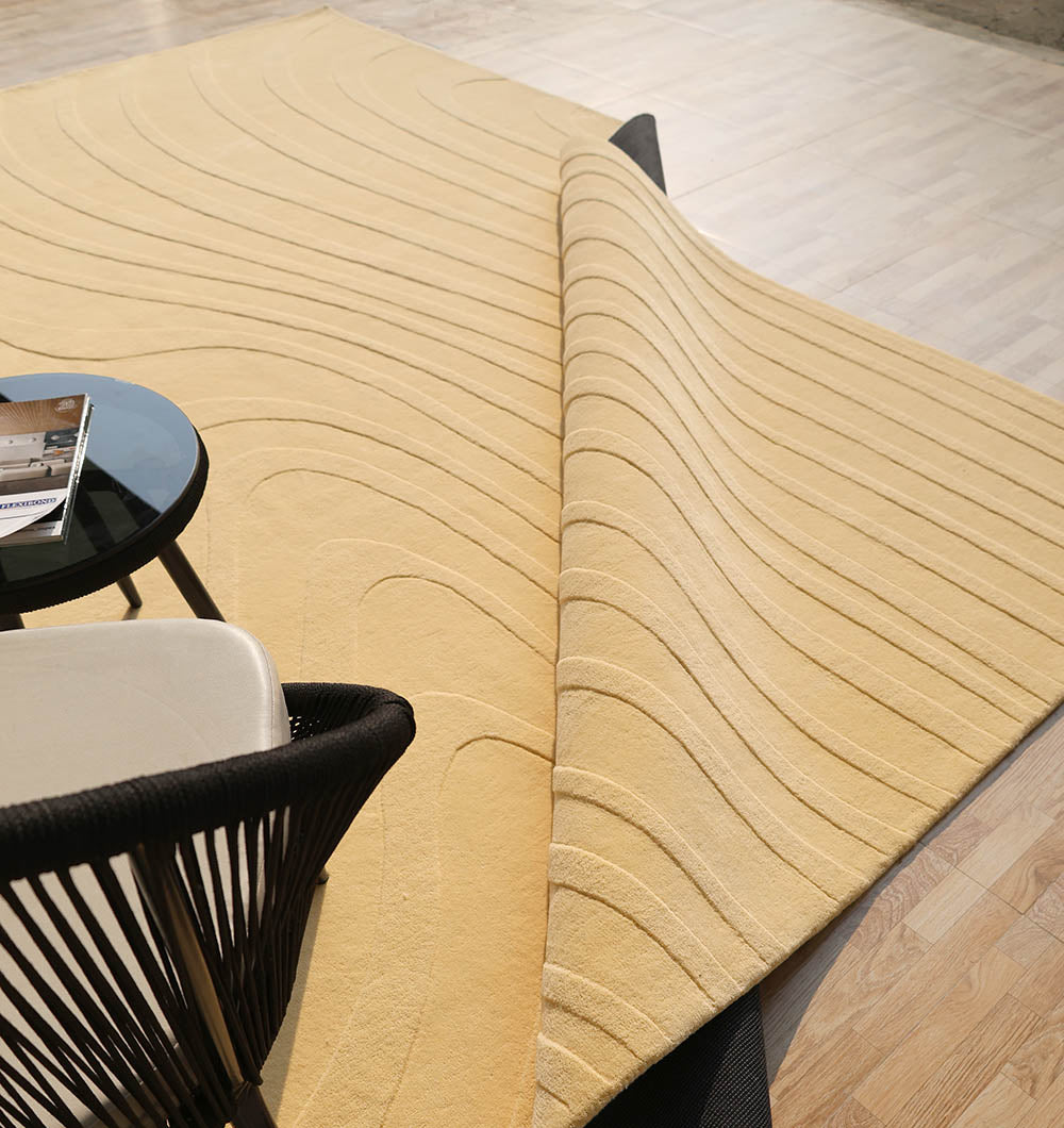 The RAMSHA HOME Minimal Wool Handmade Carpet 300x400 Yellow – HTW-117, with modern wave textures, is partially folded on a light wood floor beside a black chair and a round table holding a magazine.
