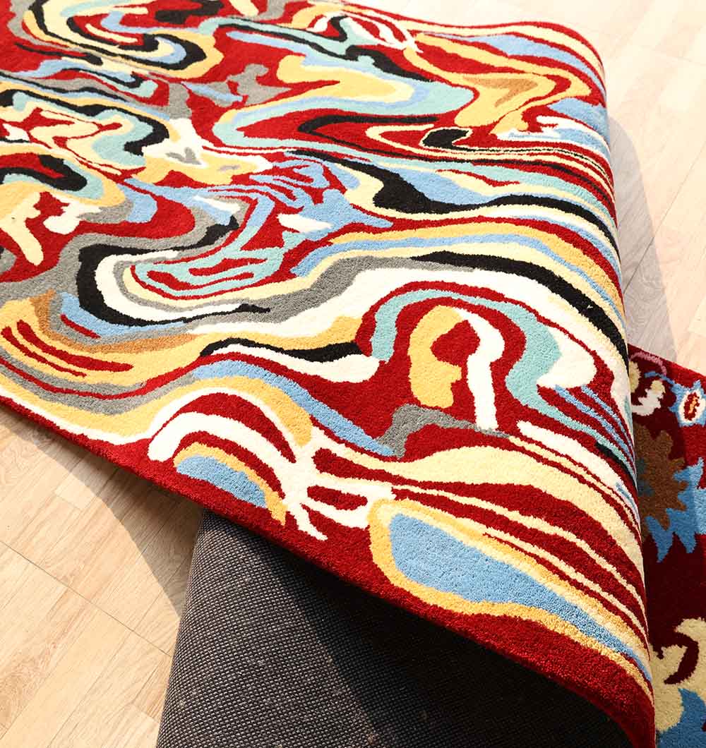 The RAMSHA HOME "Home Decor Handmade Carpet – HTW-118," a colorful, abstract-patterned hand-tufted wool rug with swirling shapes, is partially folded over a gray sofa on a light wood floor.