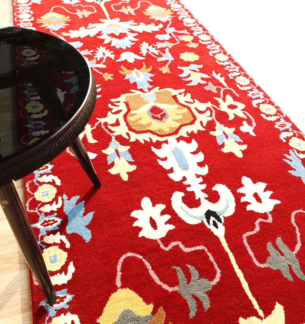 The RAMSHA HOME Handmade Carpet – HTW-118 is a vibrant red, hand-tufted rug with an intricate floral and vine pattern in white, yellow, blue, and beige. A round black table with four legs is seen on the left side of the image.