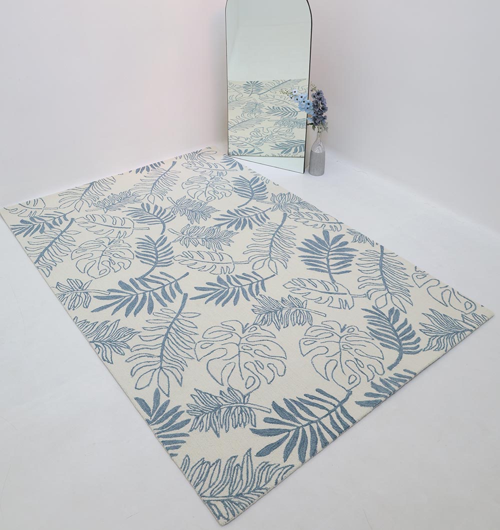 The RAMSHA HOME Home Decor Handmade Carpet – HTW-119, featuring a blue botanical pattern on a white background, is displayed on a white floor beside a mirror and a vase with blue flowers.