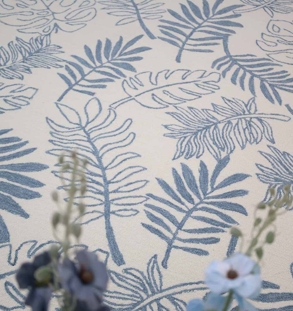 The RAMSHA HOME Home Decor Handmade Carpet – HTW-119 is a cream-colored, hand-tufted wool rug featuring a blue leaf pattern, elegantly accented by out-of-focus blue and purple flowers in the foreground.