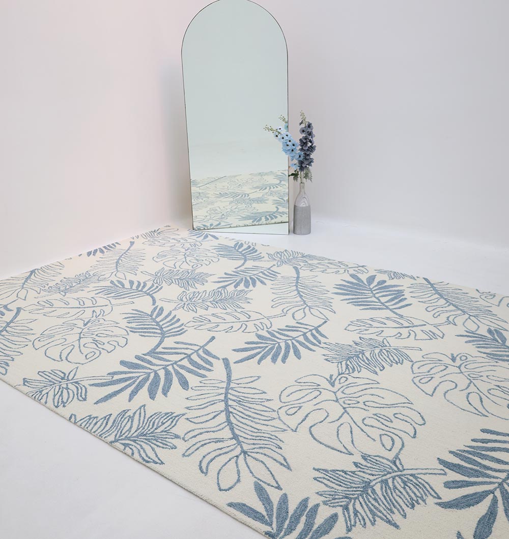 The RAMSHA HOME Handmade Carpet – HTW-119, a large cream-colored rug with blue botanical patterns, is placed on a white floor by a tall arched mirror. A gray vase with blue and white flowers beside the mirror reflects the rug’s elegant design.