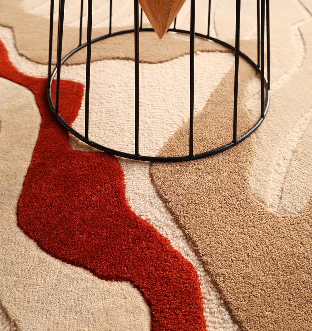 A close-up of the RAMSHA HOME Modern Handmade Carpet 300x400 Multi – HTW-120, a hand tufted wool rug with beige, tan, and red shapes. The base of a round table with black metal legs and wood center is partially visible.