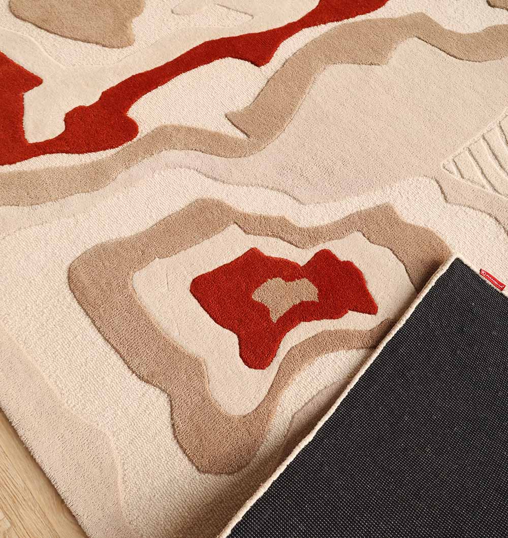 A close-up of the RAMSHA HOME Modern Handmade Carpet 300x400 Multi – HTW-120 shows its abstract layered design in beige, tan, and red. One corner is lifted to reveal a black non-slip backing underneath.