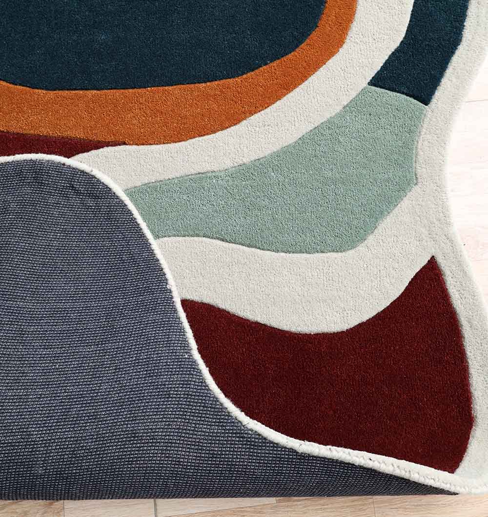 A close-up of the RAMSHA HOME Handmade Shaped Abstract Round Rug - HTW 121 (200cm), handtufted in curved blocks of dark green, orange, white, mint green, maroon, and gray, shown on a light wooden floor with one corner flipped over.