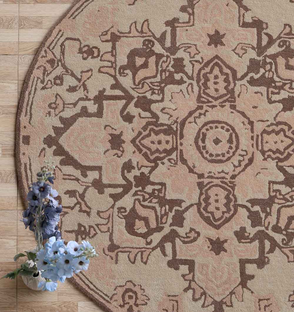 The RAMSHA HOME Handmade Cotton Round Wool Rug HTW 122 (150cm), featuring an intricate brown and beige pattern, lies on a wooden floor with a small bouquet of white and purple flowers at its lower left edge.