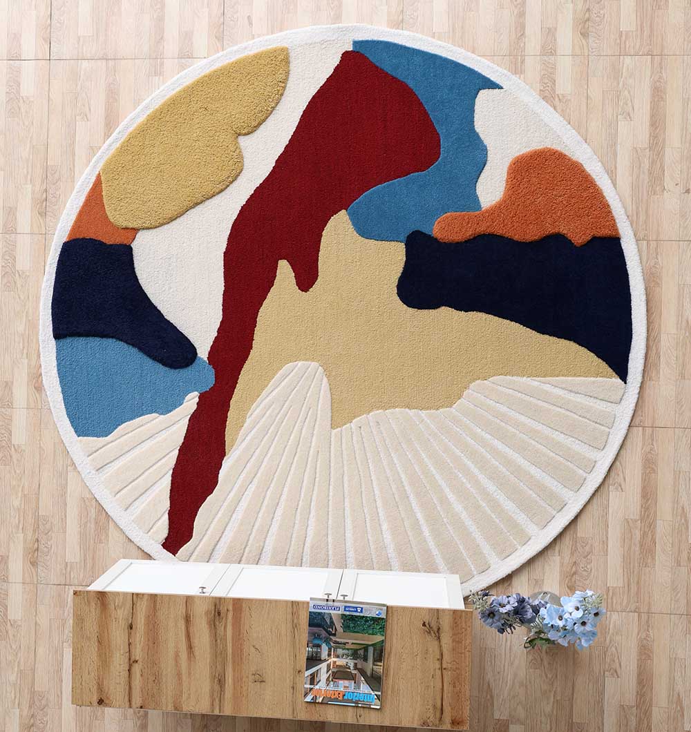 The RAMSHA HOME Handmade Wool & Cotton Abstract Round Area Rug HTW 125 (200cm), featuring bold shapes in red, blue, mustard yellow, orange, and cream, hangs on a wood-panelled wall above a wooden table with a magazine and flowers.