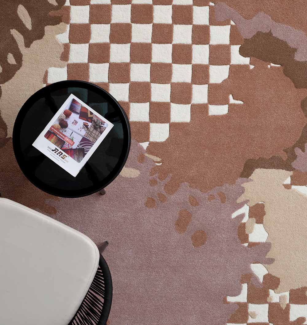 A small round black table with a magazine stands beside a light beige chair on the Handmade Multicolor Area Bespoke Rug HTW 128 (200cm) by RAMSHA HOME, featuring abstract brown and cream checkerboard and organic shapes.