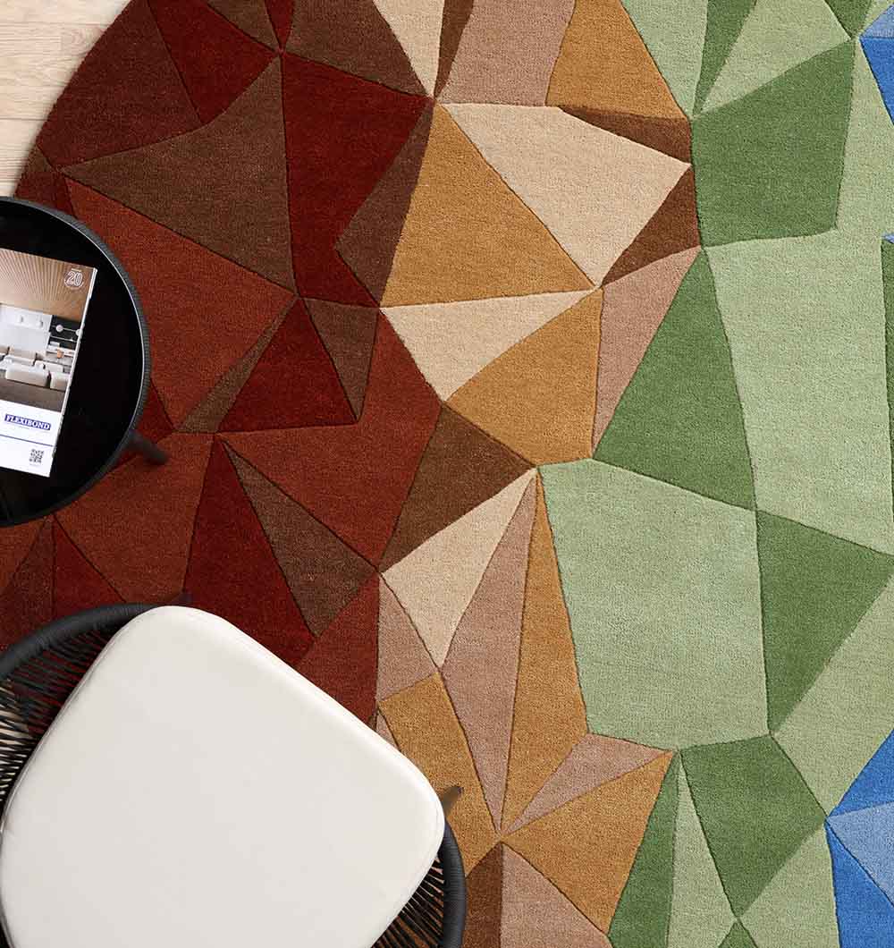 The RAMSHA HOME Multicolor Modern Geometric Round Rug HTW 130 (200cm) features vibrant green, brown, orange, and blue accents. A white chair and small round table with papers are partly seen at the image's edge.